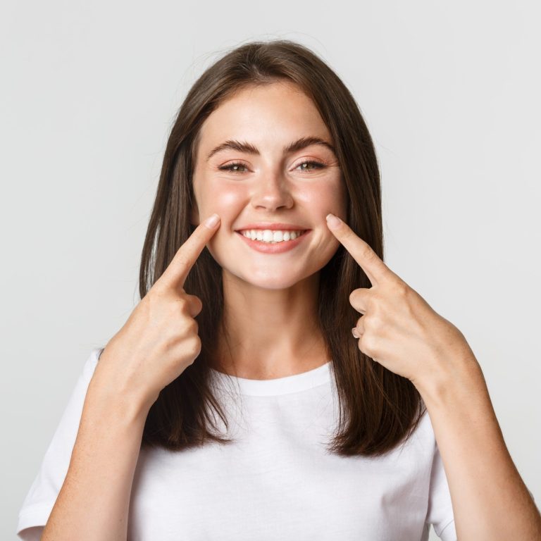 Close-up of attractive smiling young girl pointing fingers at face, poking cheeks, white background.